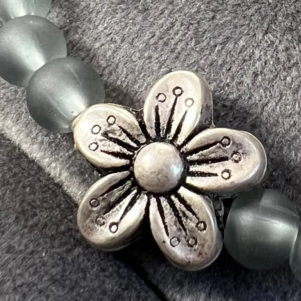 Handgjort pärlarmband i gråa pärlor med två fina silverfärgade blommor, även spännet är silverfärgat. Justerbar passform mellan 17-22 cm.. Accessoarer.
