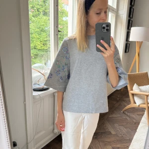 Grå tröja med broderade ärmar - By Malina. En grå tröja med broderade blommor på de vida trekvartsärmarna. Tröjan har en lös passform och är tillverkad i ett bekvämt material.