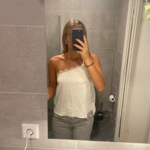 Vit offshoulder topp - En stilren vit offshoulder topp med spetsdetaljer. Toppen har en asymmetrisk design med en axelbandslös sida och en smal axelrem på andra sidan.