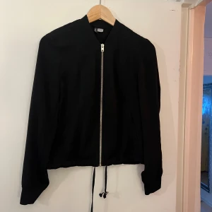 Svart jacka från H&M Divided - En svart jacka från H&M Divided med dragkedja framtill. Jackan har långa ärmar och en rak passform. Den har även en justerbar dragsko i nederkanten.