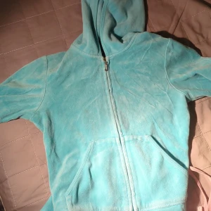 Ljusblå huvjacka från Juicy Couture - En mjuk och bekväm ljusblå huvjacka från Juicy Couture med dragkedja framtill och en stor ficka på magen. Jackan har en huva och är dekorerad med Juicy Couture-logotypen i glittriga stenar på ryggen.