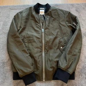 Olivgrön bomberjacka från Zara - En olivgrön bomberjacka från Zara med dragkedja framtill och ribbade muddar vid ärmslut och nederkant. Jackan har två fickor med knappar och en liten ficka på ärmen. I bra skick! 
