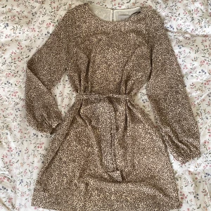 Mönstrad klänning - Supersöt mönstrad klänning med rosett i midjan! Beige med brunt och svart mönster och guld detaljer. Storlek 40 och jättebra skick💗