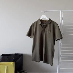 Grön Hugo Boss Passenger Polo Shirt - Helt ny med etiketten mörk grön slim fit polotröja av Hugo Boss. Oanvänd, Nypris 1075 kr. Tröjan är stilig, andningsbar och stretchig. Den har även ett CmiA-märke, som står för att förbättra levnadsförhållandena för afrikanska bomullsodlare!