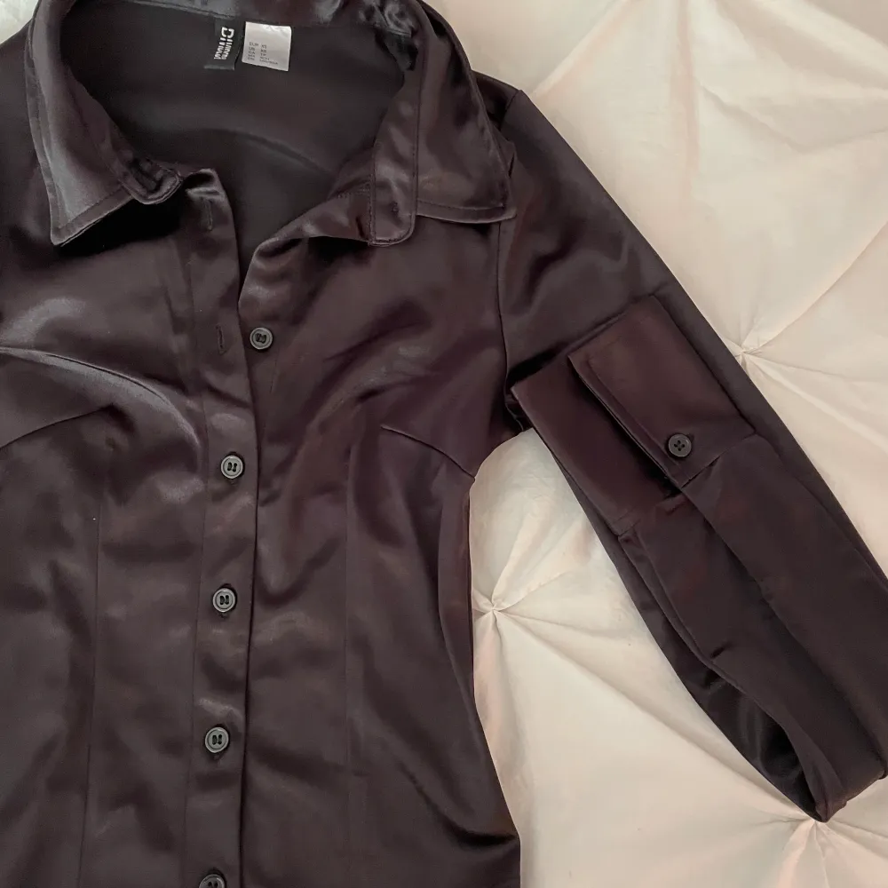 En oanvänd blus/skjorta från H&M i svart/mörkbrun nyans. Satin liknande material, storlek XS. Plagget har en ganska figurnära passform. . Blusar.