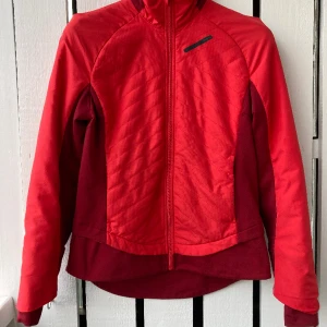 Längdskidjacka Craft - Längdskidjacka Craft, storlek M, röd och vinröd. Tunnt vadderad över bålen. Stretchiga partier i sidorna, under armarna samt nere vid höfterna. Fleeceklädd krage. Två fickor med dragkedja framtill. Liten svart fläck fram som inte gått bort i tvätten.