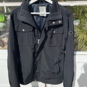 J.Lindeberg ”Bailey Poly” Field jacket  - Hej, säljer en sjukt snygg helsvart field jacka från J.Lindeberg. Köpt för 3400kr. Utrustad med både Zip samt knappar. Regntålig. Lätt att stylea till annat. Använd men fortfarande i bra skick. Storlek M. Pris kan diskuteras. Hör av dig vid intresse!