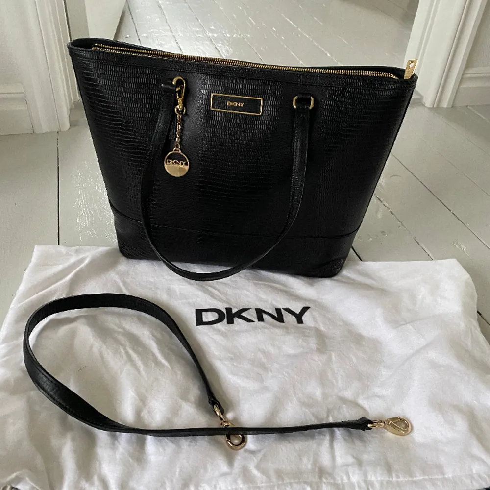 Svart DKNY handväska i skinn med lite ”ormskinnsmönster” och dragkedja. Kan både hängas på axel eller fästa rem så att man har den ”cross body”.   Helt ny, köpt för flera år sedan men aldrig använd. Dust bag medföljer. Nypris ca 2800 kr. Finns i Jönköping. Väskor.