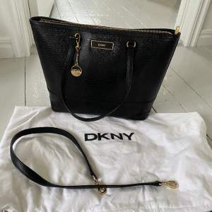 Svart DKNY handväska i skinn med lite ”ormskinnsmönster” och dragkedja. Kan både hängas på axel eller fästa rem så att man har den ”cross body”.   Helt ny, köpt för flera år sedan men aldrig använd. Dust bag medföljer. Nypris ca 2800 kr. Finns i Jönköping