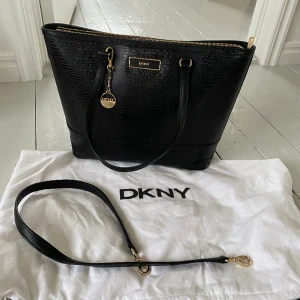 Helt ny DKNY handväska  - Svart DKNY handväska i skinn med lite ”ormskinnsmönster” och dragkedja. Kan både hängas på axel eller fästa rem så att man har den ”cross body”.   Helt ny, köpt för flera år sedan men aldrig använd. Dust bag medföljer. Nypris ca 2800 kr. Finns i Jönköping