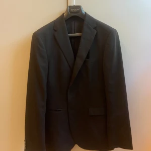 Svart Tiger of Sweden kostym i storlek 100 (kort 50) - Svart Tiger of Sweden kostym i storlek 100, vilket motsvarar en kortare 50. Färgkod 050.  Modellen på kavaj heter STYLE HENRIE 2B. Ärmlängd 64cm. 52cm över bröstet då kavajen är knäppt (från armhåla till armhåla). Tillhörande kostymbyxa ingår!  