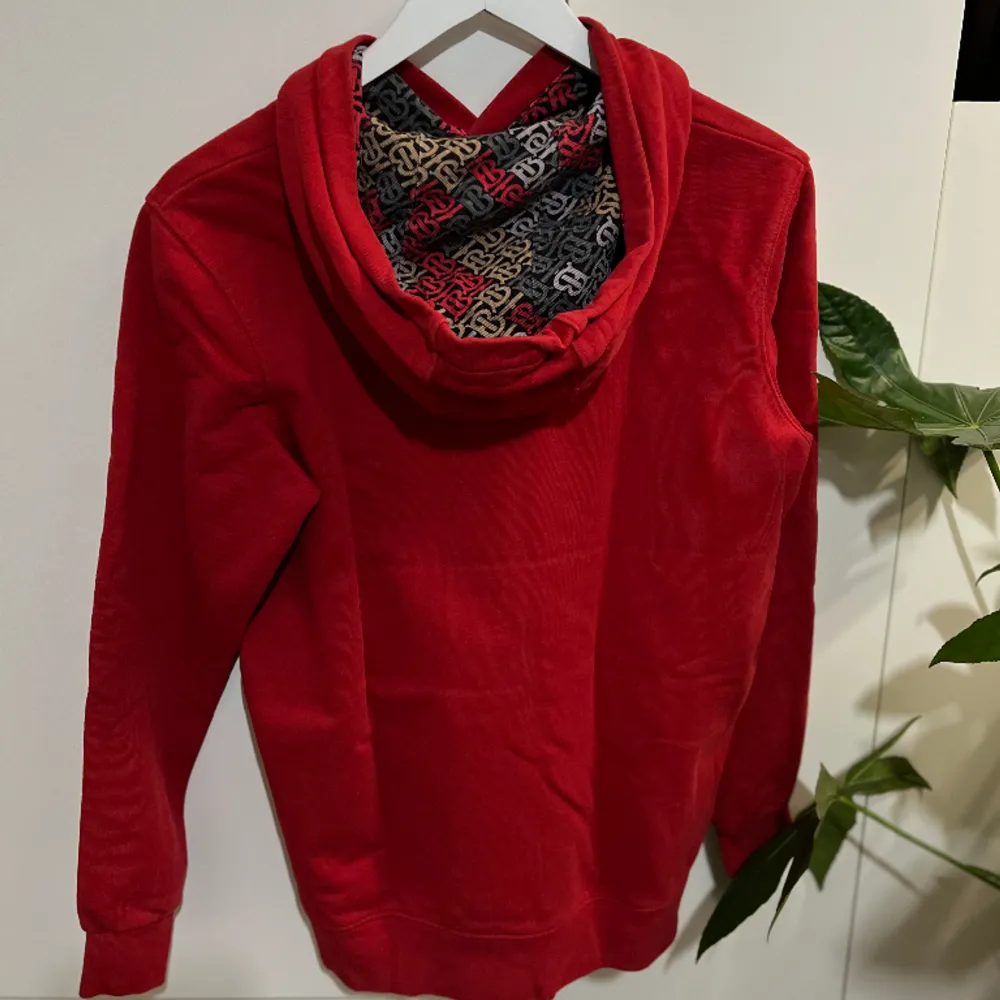 En Burberry Hoodie , i en härlig röd färg, väntar på att bli en del av din garderob! Denna klassiska och trendiga huvtröja är perfekt för att ge din outfit en touch av lyx och komfort.  Axelbred:42cm Längd: 68cm. Hoodies.