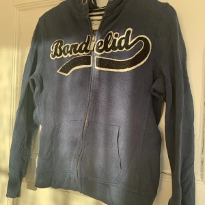 Mörkblå Zipup  - En mörkblå Zipup med ”tryck” på. Den är i storlek M men sitter mer som S, och kommer från Bondelid. Har inga hål/fläckar men är välanvänd och är därför lite nopprig på texten.