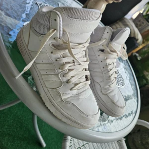 Vita sneakers från Adidas - Ett par vita sneakers från Adidas med snörning och hög ankel. Skorna har en robust sula och är tillverkade i syntetmaterial med mesh-detaljer för ökad andningsförmåga. Adidas-logotypen syns tydligt på baksidan av skorna.