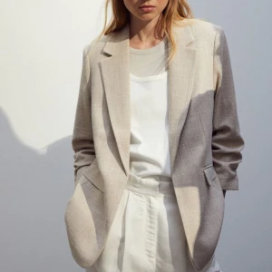 Kavaj - Figurnära kavaj från H&M. Går inte längre att köpa. Inga skador eller skavanker. Är i vävd kvalitet av viskosblandning. Har axelvaddar, framfickor med lock, och rynkad trekvartslång ärm.  Har ingen knäppning. 