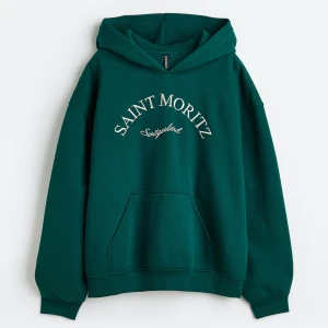 Grön hoodie från Divided - En bekväm grön hoodie från Divided med texten 'Saint Moritz' tryckt på framsidan. Hoodien har en känguruficka och ribbstickade muddar vid ärmslut och nederkant.