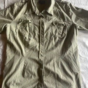 Olivgrön skjorta från GR Design - En olivgrön skjorta från GR Design med korta ärmar och två bröstfickor med lock. Skjortan har en klassisk krage och knäppning framtill.