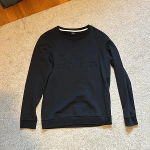 Svart tröja från Hugo Boss - (PERFEKT SKICK!!!) En mörkblå långärmad tröja från Hugo Boss med en rund halsringning. Tröjan har en diskret logotyp i samma färg på framsidan. Tröjan har endast blivit använd ett fått antal gånger och det är inte möjligt att se att den är använd.