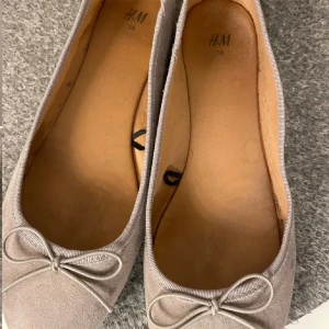 Beige ballerinaskor från H&M - Ett par beige ballerinaskor från H&M i storlek 38. Skorna har en rosett på tån och är tillverkade i ett mjukt material.