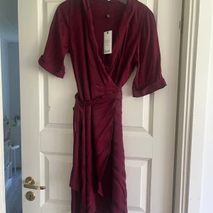 Burgundy Stripe Midiklänning från Dry Lake - En elegant burgundy midiklänning från Dry Lake med omlott-design och knytband i midjan. Klänningen har trekvartsärmar och är tillverkad av 52% rayon och 48% viskos.