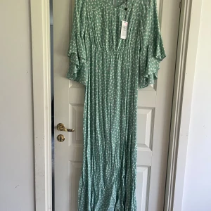 Grön långklänning med vita mönster - En grön långklänning med vita mönster. Klänningen har vida ärmar och ett löst passande snitt. Den är tillverkad i ett lätt och luftigt material, perfekt för varmare väder.