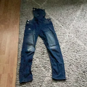Blå jeansbyxdress med slitningar - Säljer en snygg blå jeansbyxdress med slitningar på knäna. Den har justerbara axelband och knappar på sidorna för en perfekt passform. Perfekt för en avslappnad och cool look!