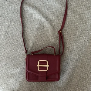 Vinröd axelväska med gulddetaljer - Säljer en snygg vinröd axelväska i skinn med justerbar axelrem och ett handtag. Väskan har en stor guldfärgad spänne på framsidan som detalj💕