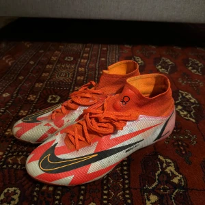 Nike Mercurial fotbollsskor - Säljer ett par Nike Mercurial fotbollsskor i nyskick. Skorna är i en cool orange och vit färgkombination med svarta detaljer och snörning. De har en tight passform och är perfekta för fotbollsträning och matcher. Sulan har en bra greppdesign för att ge bästa möjliga fäste på planen.