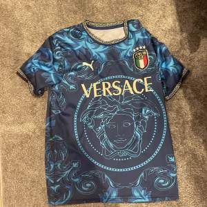 Säljer en snygg blå fotbollströja från Versace med ett coolt mönster i olika nyanser av blått. Tröjan har korta ärmar och detaljer i guld på kragen och ärmsluten. Den har också Italiens landslagsemblem på bröstet och Versace-loggan i guld på framsidan. Perfekt för dig som vill sticka ut på planen!