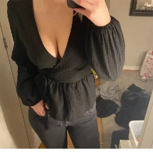 Svart omlottopp med knytband - Säljer en snygg svart omlottopp i storlek 36. Toppen har långa ärmar och ett knytband i midjan som ger en fin passform. Perfekt för både vardag och fest!