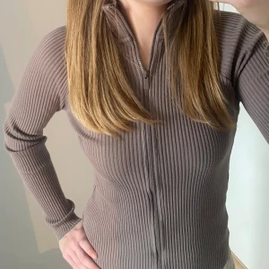 RIBSTICKAD TRÖJA - Ribstickad tröja med dragkedja. Luftigt material så man kan ha den som träningströja. Färgen är en blandning mellan lila, brun och grå. Skriv ifall du har några funderingar ang plagget.😊