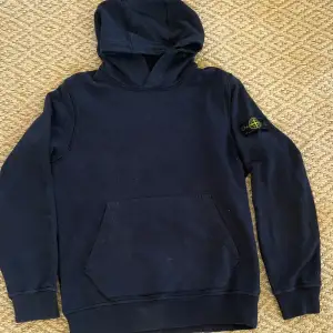 Snygg Mörkblå Stone Island-hoodie i toppskick! Passar perfekt för dig som är 165-170 cm lång. Ursprungligen köpt för 2200 kr, nu tillgänglig för endast 1400 kr (priset kan diskuteras). Nästan oanvänd och äkta med QR-kod och extra knapp inkluderad.