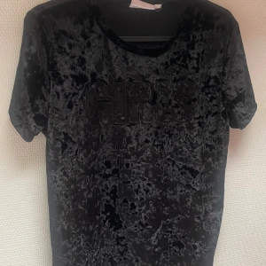 Svart t-shirt från KappAhl - En svart t-shirt från KappAhl med texten 'GIRLS' i relief på framsidan. T-shirten har korta ärmar och är tillverkad i ett glansigt material.