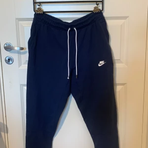 Mörkblå mjukisbyxor från Nike - Säljer ett par mörkblå mjukisbyxor från Nike i storlek M. Byxorna har en elastisk midja med vit snörning och en liten vit Nike-logga på vänster ben. De är superbekväma och perfekta för chill eller träning. Byxorna har också en bakficka. Köp dem nu eller ge ett bud! 💙