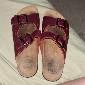 Röda sandaler från Scholl - Säljer ett par röda sandaler från Scholl i storlek 40. De har två justerbara remmar med svarta spännen och en bekväm fotbädd. Sandalerna är i bra skick och perfekta för sommaren.