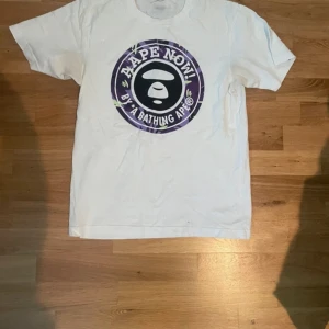 Vit t-shirt från Aape by A Bathing Ape - En vit t-shirt från Aape by A Bathing Ape med ett stort, runt tryck i lila och svart på framsidan. T-shirten är tillverkad av 100% bomull och har korta ärmar. storleken är S👍👍
