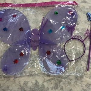 Fjärilsvingar med diadem och trollstav - Säljer ett set med fjärilsvingar, diadem och trollstav. Finns i flera färger: lila, rosa, mörkrosa och röd. Vingarna är dekorerade med glittriga blommor och setet är perfekt för fest eller utklädnad. Vingarna är gjorda av plast och diademet har små fjärilar på sig. 75kr/ paket. sammafrakt 