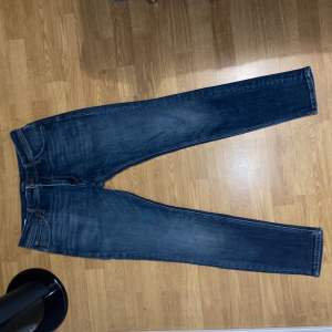 Ett par blå skinny fit jeans från Jack & Jones, modell Liam. Jeansen har en klassisk femficksdesign och är tillverkade i denim med orange sömmar.