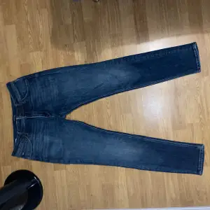 Ett par blå skinny fit jeans från Jack & Jones, modell Liam. Jeansen har en klassisk femficksdesign och är tillverkade i denim med orange sömmar.