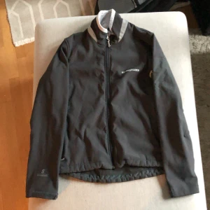 Svart vindjacka från Henri Lloyd - En svart vindjacka från Henri Lloyd med dragkedja framtill. Jackan har långa ärmar och en hög krage med vit och grå detalj. Den är tillverkad i ett slitstarkt material och har en normal passform. Nästan aldrig använd!