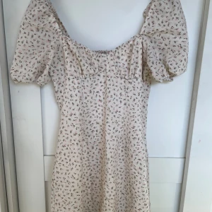 Blommig klänning - Klänning med små rosa blommor. Ryggen är stretchig genom rynkning som man ser på tredje bilden. På vänster sida sitter en dragkedja under ärmen. 