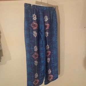 Blommiga byxor från Ida Sjöstedt för Monki - Ett par blå byxor med blommönster från Ida Sjöstedt för Monki. Byxorna har ett löst passform och är dekorerade med rosa och vita blommor.