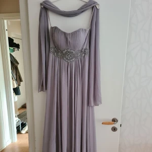 Klänning - Fin axelbandslös balklänning i grå/lila färg. Följer med en matchande bolero/tyg att ha över axlarna. Storlek S. Har en liten skada som syns inringad på ena bilden. Går nog att laga lätt med något stygn.