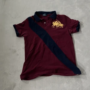 Vinröd pikétröja från Polo Ralph Lauren - En vinröd pikétröja från Polo Ralph Lauren med en mörkblå diagonal rand och ett broderat gult polospelmotiv på bröstet. Tröjan har korta ärmar och en klassisk krage med två knappar.