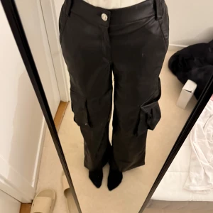 Svarta cargobyxor i skinnimitation - Säljer ett par svarta cargobyxor i skinnimitation med flera fickor på sidorna och raka ben. Byxorna har normal passform och stängs med knapp och dragkedja. Perfekta för en cool och trendig look.