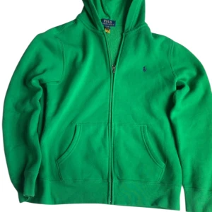 Grön hoodie från Polo Ralph Lauren - Säljer en grön hoodie från Polo Ralph Lauren med dragkedja och två fickor framtill. Klassisk broderad logga på bröstet. Perfekt för en avslappnad stil och enkel att matcha med jeans eller joggers.