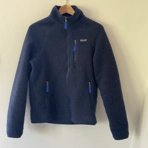 Mörkblå fleecejacka från Patagonia - Mysig mörkblå fleecejacka från Patagonia med hög krage och tre fickor med blå dragkedjor. Perfekt för kyliga dagar eller sena sommarkvällar. 