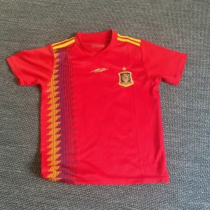 Spansk fotbollströja med gult mönster - Röd kortärmad fotbollströja från Luanvi med Spaniens landslagsemblem på bröstet. Tröjan har gula och blå grafiska detaljer längs ena sidan samt gula ränder på axlarna. Perfekt för fotbollsfans!