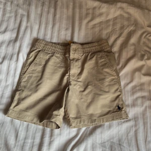 Beige shorts från Polo Ralph Lauren - Snygga beige shorts från Polo Ralph Lauren med elastisk midja och sidofickor. Klassisk design med broderad logga framtill och två bakfickor med knapp. Perfekta för varma dagar och enkel att matcha med det mesta.