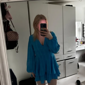 Blå kort jumpsuit med volang och v-ringning - Supersöt blå jumpsuit/klänning med v-ringning, långa ärmar och volangdetaljer. Klänningen har markerad midja och omlottkänsla med knyt i midjan. Perfekt för dig som vill sticka ut med färg och stil.